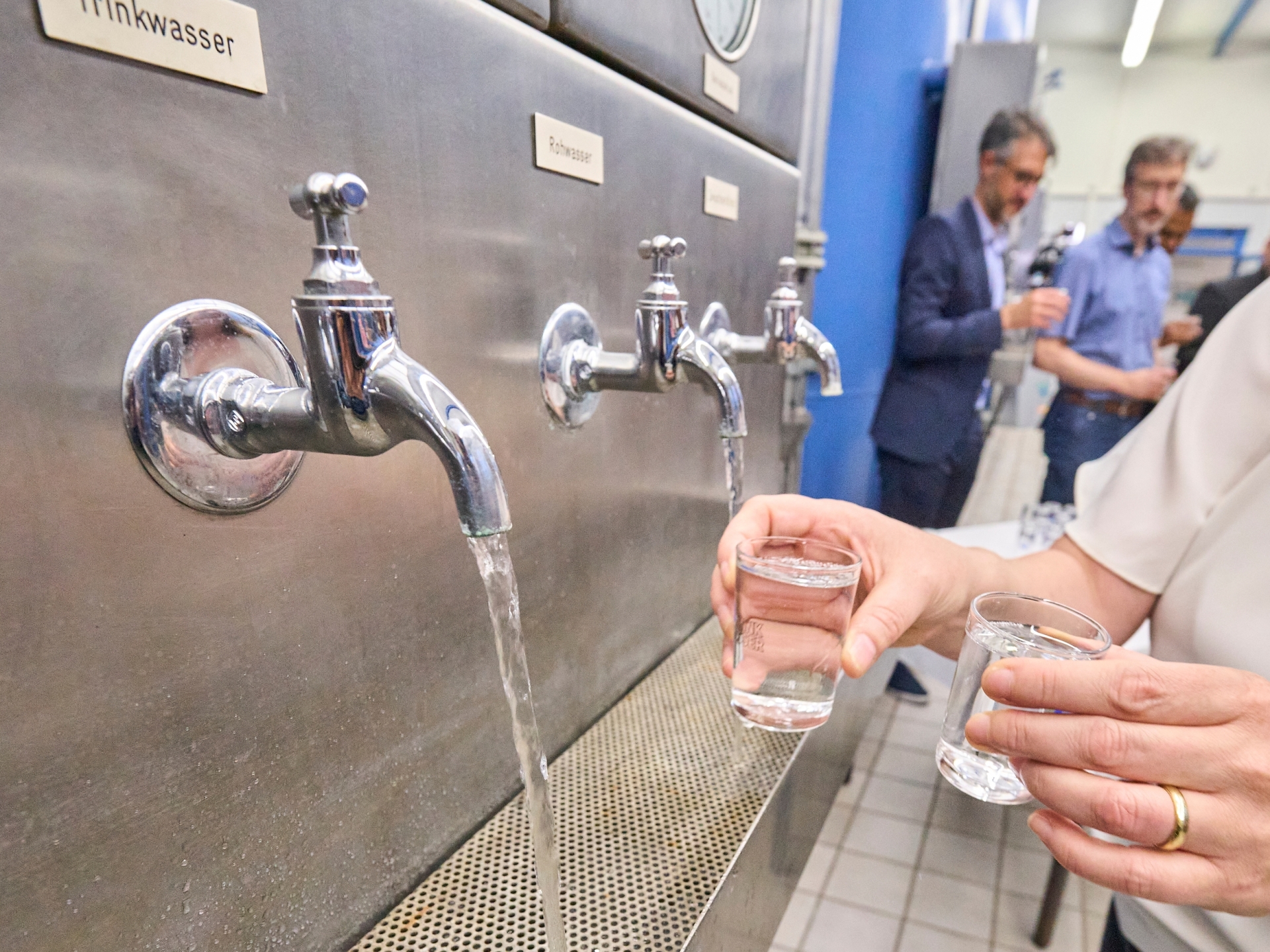 Mehrere Wasserhähne an einer Edelstahlstation geben Trinkwasser aus, während eine Person zwei kleine Gläser mit Wasser befüllt. Im Hintergrund stehen weitere Personen in einem technischen Raum.