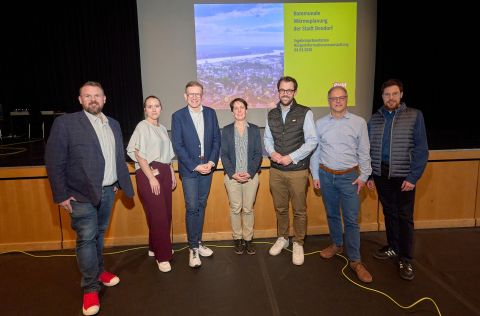 Eine Gruppe von sieben Personen steht im vorderen Bereich einer Bühne in der Bendorfer Stadthalle. Hinter ihnen ist eine große Leinwand mit einer Präsentationsfolie zur kommunalen Wärmeplanung der Stadt Bendorf zu sehen. Die Personen stehen nebeneinander und repräsentieren die beteiligten Institutionen der Veranstaltung.