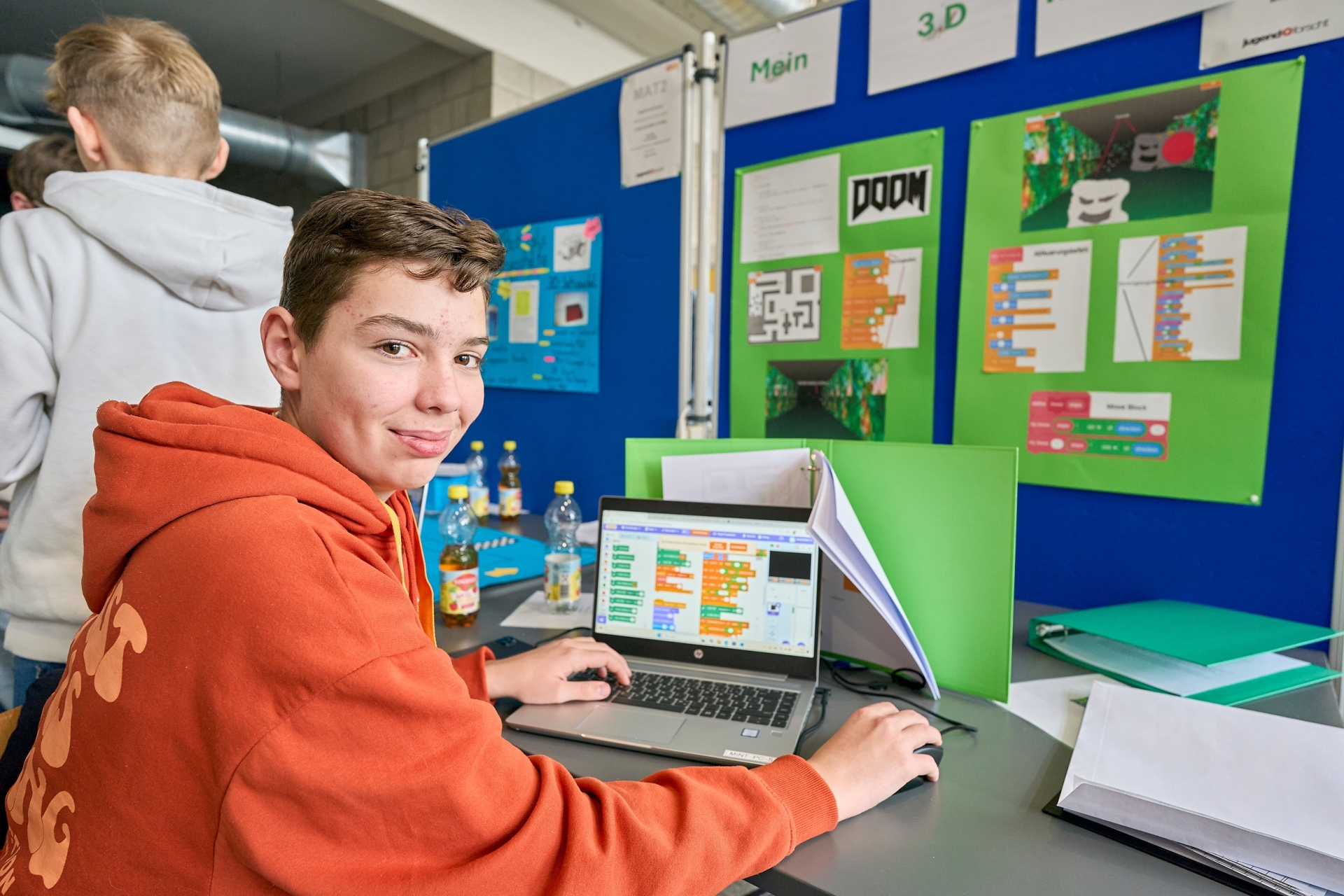 An einem „Jugend forscht“-Stand arbeitet ein Teilnehmender an einem Laptop mit einer visuell programmierten 3D‑Irrgarten‑Software; hinter ihm hängen Poster mit Screenshots und Projektinformationen.