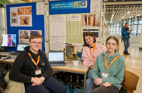 Jugend forscht 2026 - Regionalwettbewerb Koblenz Drei Teilnehmende sitzen an ihrem „Jugend forscht“-Stand und zeigen ein automatisches Bewässerungssystem mit Sensorik und Pflanzenmodell vor einer Stellwand mit Projektpostern.