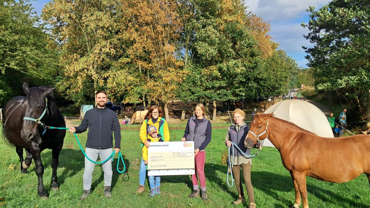 Mehrere Personen stehen gemeinsam mit zwei Pferden auf einer Wiese vor einem Zelt und halten einen symbolischen Spendenscheck der evm-Gruppe über 1.000 Euro. Die Spende kommt dem Verein Pegasus e.V. zugute, der sich unter anderem für pferdegestützte Angebote zur Förderung von Kindern, Jugendlichen und Erwachsenen engagiert.