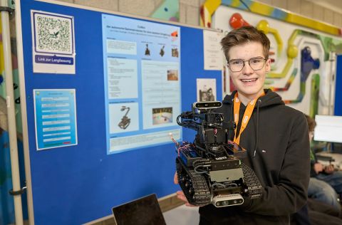 Jugend forscht 2026 - Regionalwettbewerb Koblenz Eine teilnehmende Person des Wettbewerbs „Jugend forscht“ hält einen selbst konstruierten Roboter mit Greifarm und Kettenantrieb in den Händen. Im Hintergrund befindet sich ein Präsentationsstand mit einem wissenschaftlichen Poster zum Projekt „Colbot“, das Aufbau, Funktionsweise und Entwicklungsschritte des Roboters zeigt. Daneben hängen zusätzliche Informationstafeln und ein QR-Code zur weiterführenden Projektbeschreibung.