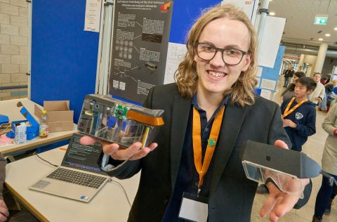 Teilnehmende Person zeigt am Stand des Wettbewerbs ein technisches Projekt: zwei Prototypen einer Datenbrille, darunter ein durchsichtiges Gerät mit sichtbarer Elektronik und ein kompaktes schwarzes Modul. Im Hintergrund sind ein Laptop, ein Projektposter und weitere Besucherinnen und Besucher der Veranstaltung zu sehen.