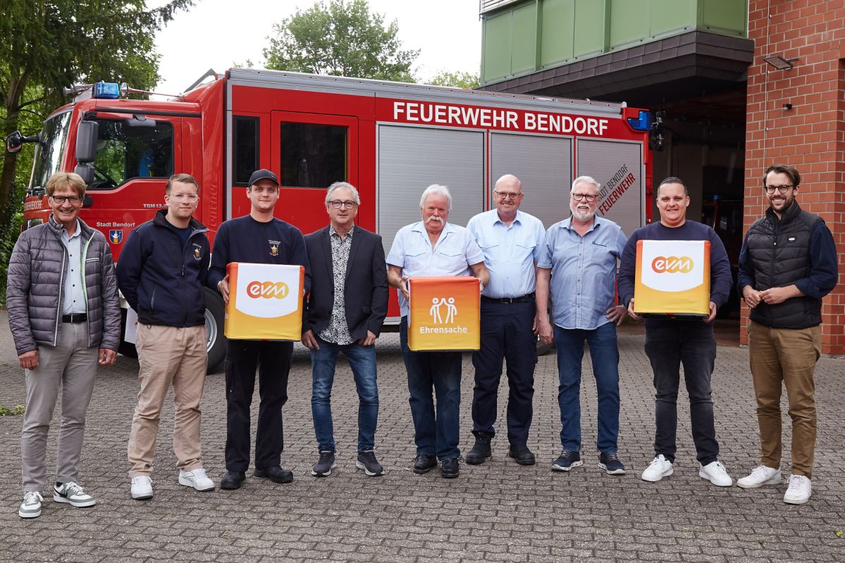 evm-Ehrensache Berthold Nick von der evm und der Erste Beigeordnete Peter Schneider stehen mit Vertreterinnen und Vertretern der Feuerwehr‑Fördervereine vor einem Feuerwehrfahrzeug und halten „evm‑Ehrensache“-Spendenboxen bei der Übergabe.