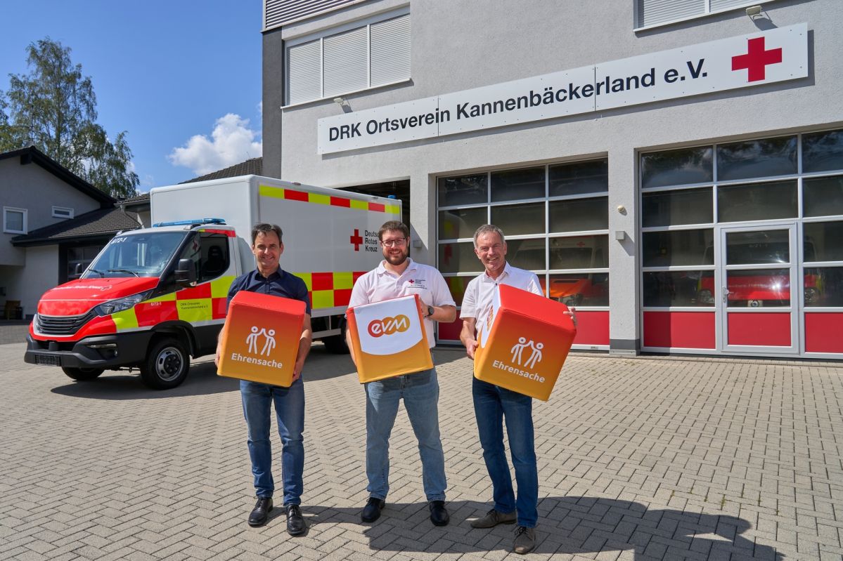 evm-Ehrensache Drei Personen stehen vor dem Gebäude des DRK Ortsvereins Kannenbäckerland und halten „evm‑Ehrensache“-Spendenboxen. Im Hintergrund sind ein Einsatzfahrzeug des Deutschen Roten Kreuzes sowie die Fahrzeughalle des Ortsvereins zu sehen. Norbert Rausch von der evm, Bürgermeister Marco Weißer und DRK‑Vertreter Torsten Michalsky präsentieren die Spende.