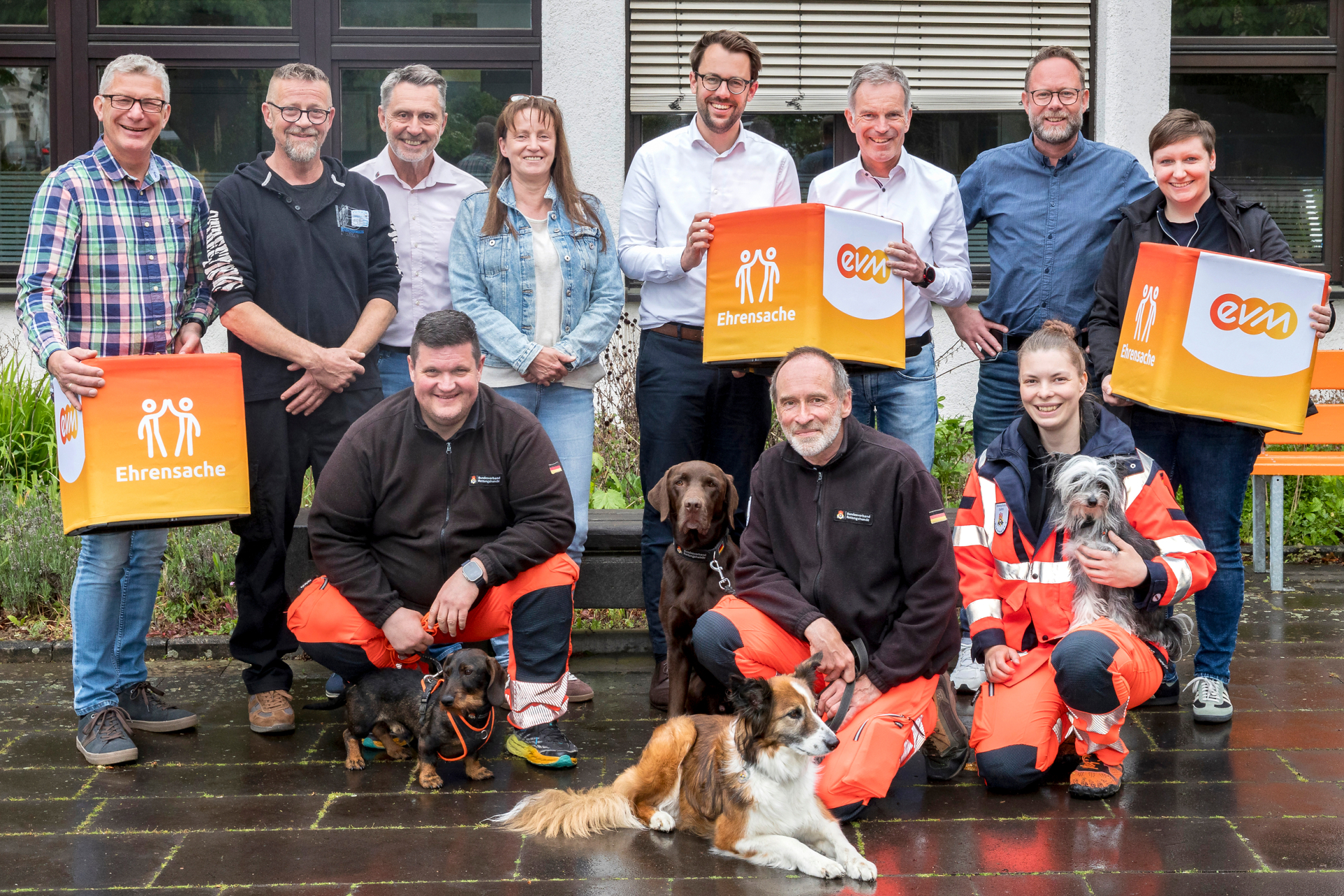 Norbert Rausch und Hubertus Hacke‑Dietze von der evm sowie Verbandsbürgermeister Sven Schür stehen mit Vertreterinnen und Vertretern mehrerer Vereine, darunter eine Rettungshundestaffel mit Hunden, bei der Übergabe der „evm‑Ehrensache“-Spende vor einem Gebäude.