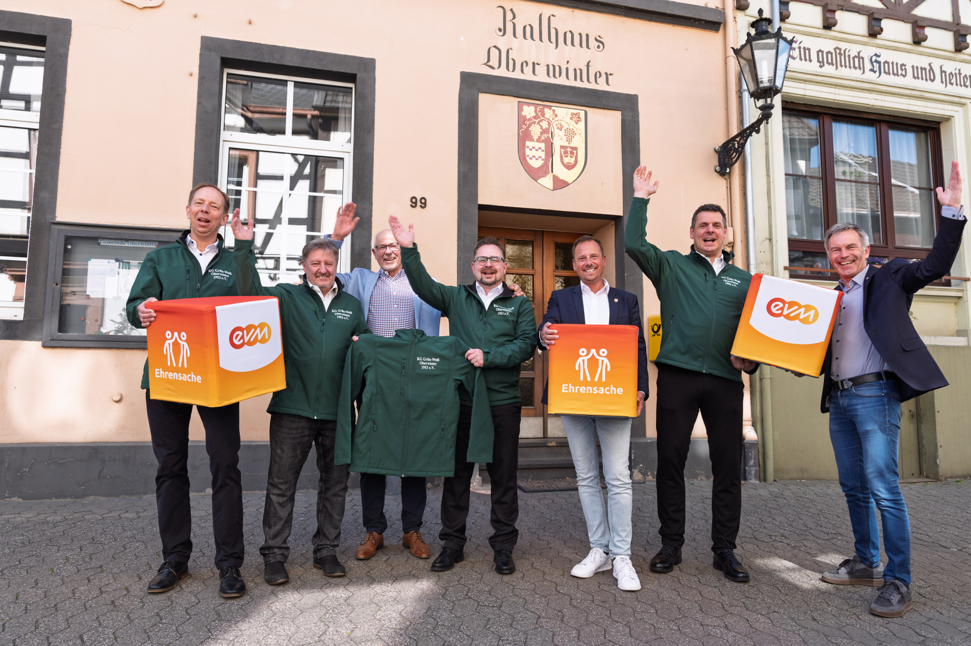 Norbert Rausch von der evm, Bürgermeister Björn Ingendahl, Ortsvorsteher Jürgen Walbröl sowie Vertreter der KG Grün‑Weiß Oberwinter stehen vor dem Rathaus Oberwinter und halten „evm‑Ehrensache“-Spendenboxen bei der symbolischen Übergabe.