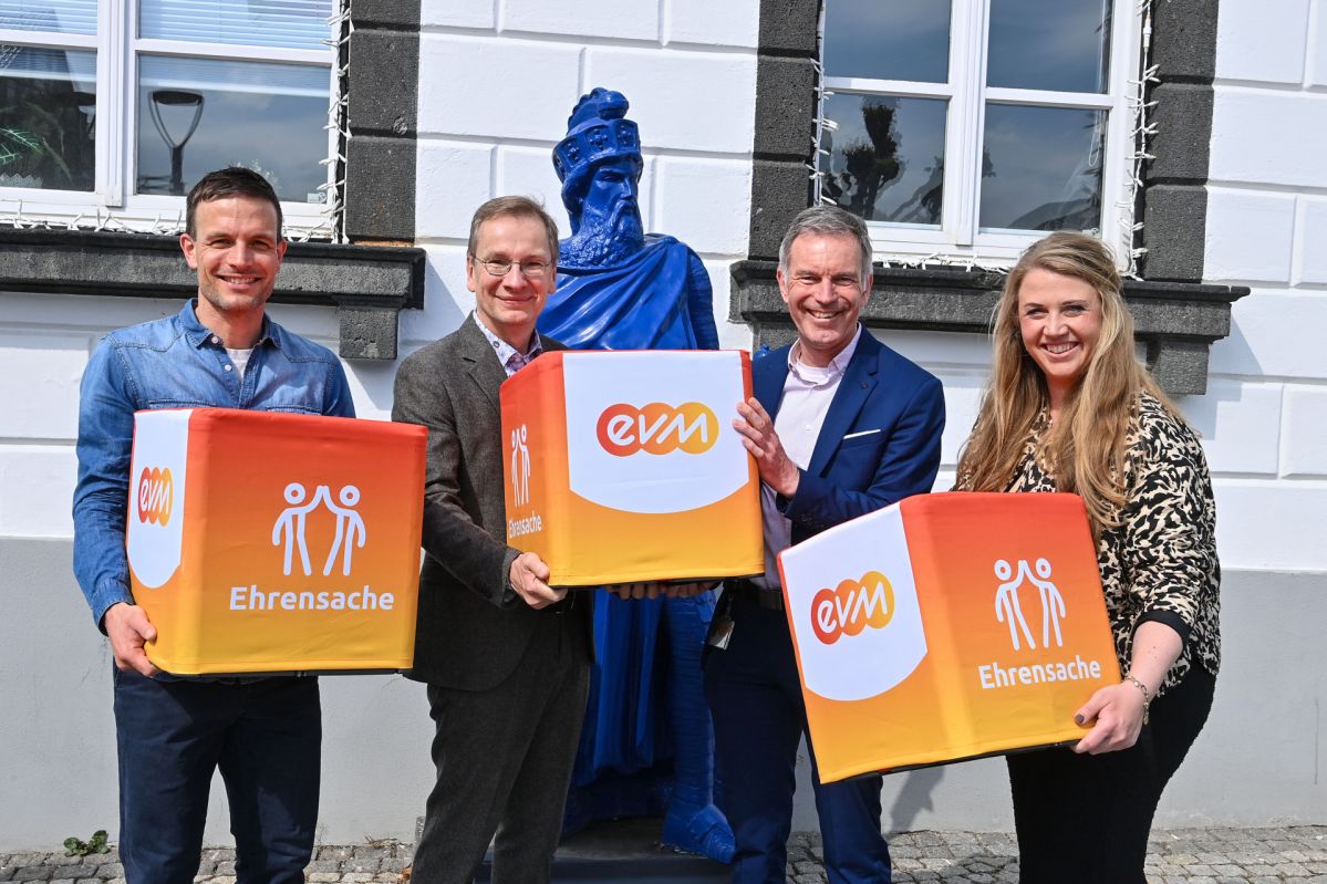 Norbert Rausch von der evm und Sinzigs Bürgermeister Andreas Geron stehen gemeinsam mit Vertreterinnen und Vertretern der Kitas Max und Storchennest vor einem Gebäude in Sinzig und halten Spendenwürfel des Programms „evm‑Ehrensache“ zur Übergabe von insgesamt 2.000 Euro.