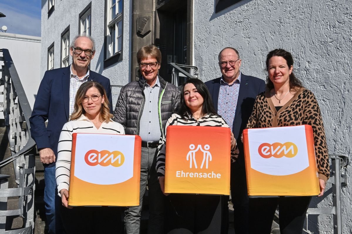 Berthold Nick von der evm und Bürgermeister Guido Nisius stehen vor einem Gebäude mit Vertreterinnen und Vertretern der Fördervereine aus Adenau und halten „evm‑Ehrensache“-Spendenboxen.