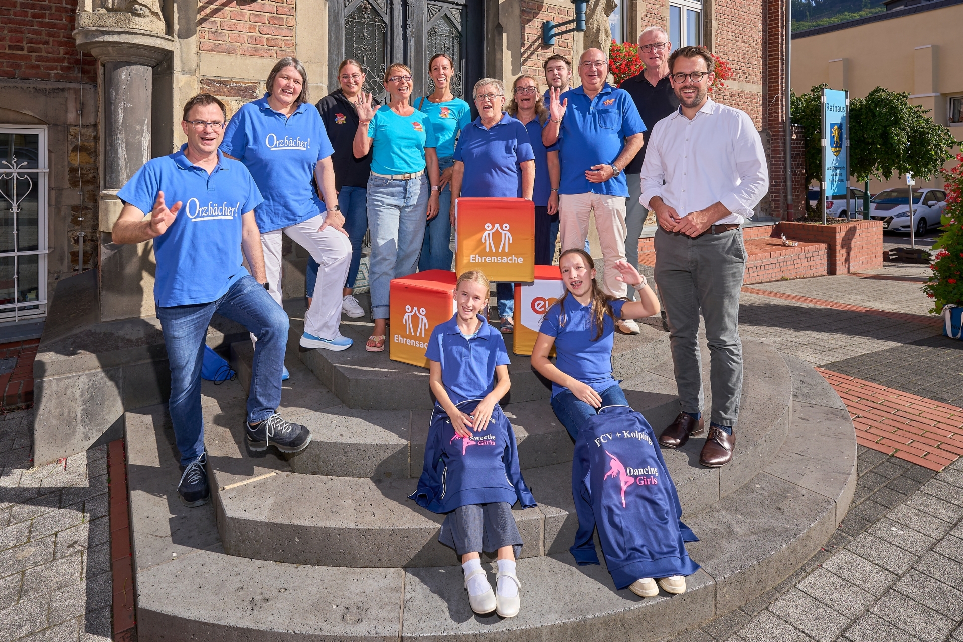 Mitglieder mehrerer Karnevalsvereine aus der Verbandsgemeinde Bad Ems‑Nassau stehen auf den Stufen vor dem Rathaus und präsentieren gemeinsam mit Kindern die „evm‑Ehrensache“-Spendenboxen. Mit dabei sind Hubertus Hacke-Dietze von der evm, Bürgermeister Uwe Bruchhäuser sowie Vertreterinnen und Vertreter des Möhnenvereins „Immer dabei“ e.V. Arzbach, des Karnevalsvereins „Blau‑Weiß“ Arzbach e.V., des Fachbacher Carnevals Vereins und der Bad Emser Karnevalsgesellschaft.