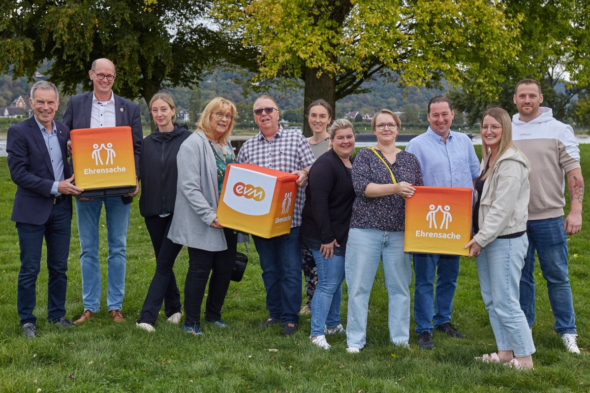 evm-Ehrensache Bad Hönningen Bürgerverein Bad Hönningen e.V., Förderverein ProJu e.V., Casa Vivida e.V., Casa Feliz e.V., Knirps e.V. und der Wild Förderverein Casa Natura e.V. stehen gemeinsam mit Norbert Rausch von der evm und Bürgermeister Jan Ermtraud auf einer Grünfläche am Rhein. Die Vertreterinnen und Vertreter der Vereine präsentieren die „evm‑Ehrensache“-Spendenboxen bei der symbolischen Spendenübergabe.