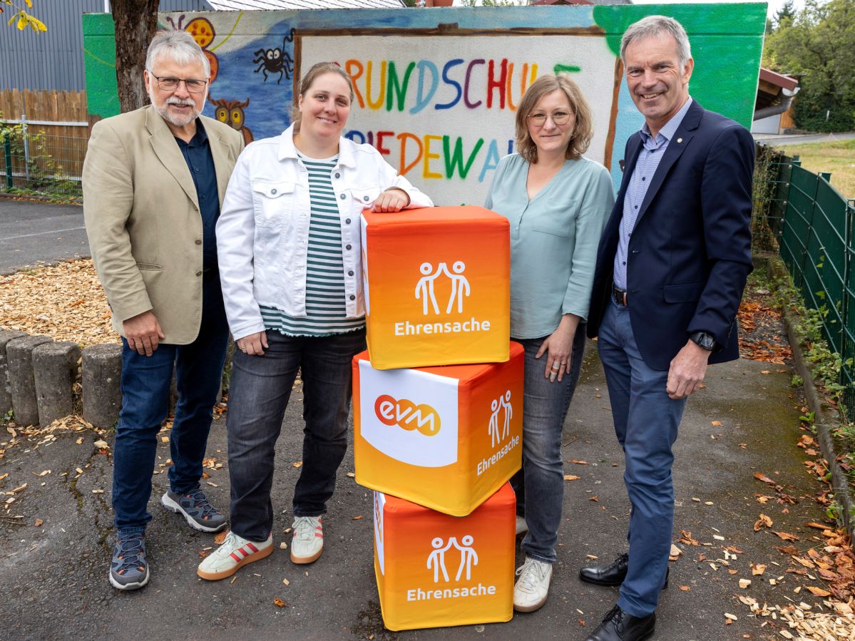 evm-Ehrensache Vier Personen stehen vor einer bunt bemalten Wand der Grundschule Friedewald. Vor ihnen sind mehrere „evm‑Ehrensache“-Spendenboxen gestapelt. Norbert Rausch von der evm, Helmut Stühn, Bürgermeister der Verbandsgemeinde Daaden‑Herdorf, sowie Vertreterinnen des Fördervereins präsentieren die Spende für die geplante Zirkusprojektwoche der Grundschule.