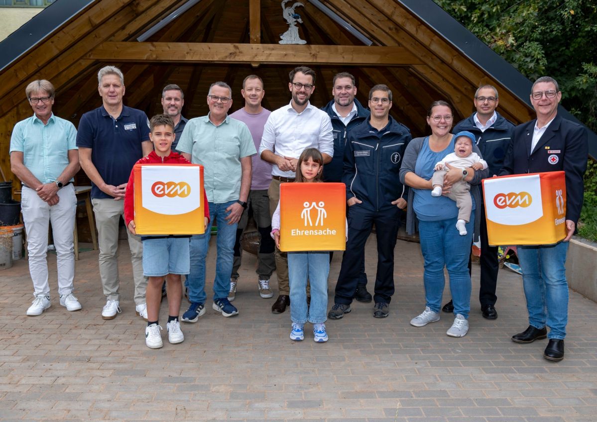 evm-Ehrensache Mehrere Vertreterinnen und Vertreter von Vereinen und Organisationen stehen vor einem hölzernen Pavillon. Im Vordergrund halten zwei Kinder „evm‑Ehrensache“-Spendenboxen. Gemeinsam mit Hubertus Hacke-Dietze und Berthold Nick von der evm sowie Hans Peter Böffgen, Bürgermeister der Verbandsgemeinde Gerolstein, präsentieren sie die Spende für die Jugendarbeit in der Region.