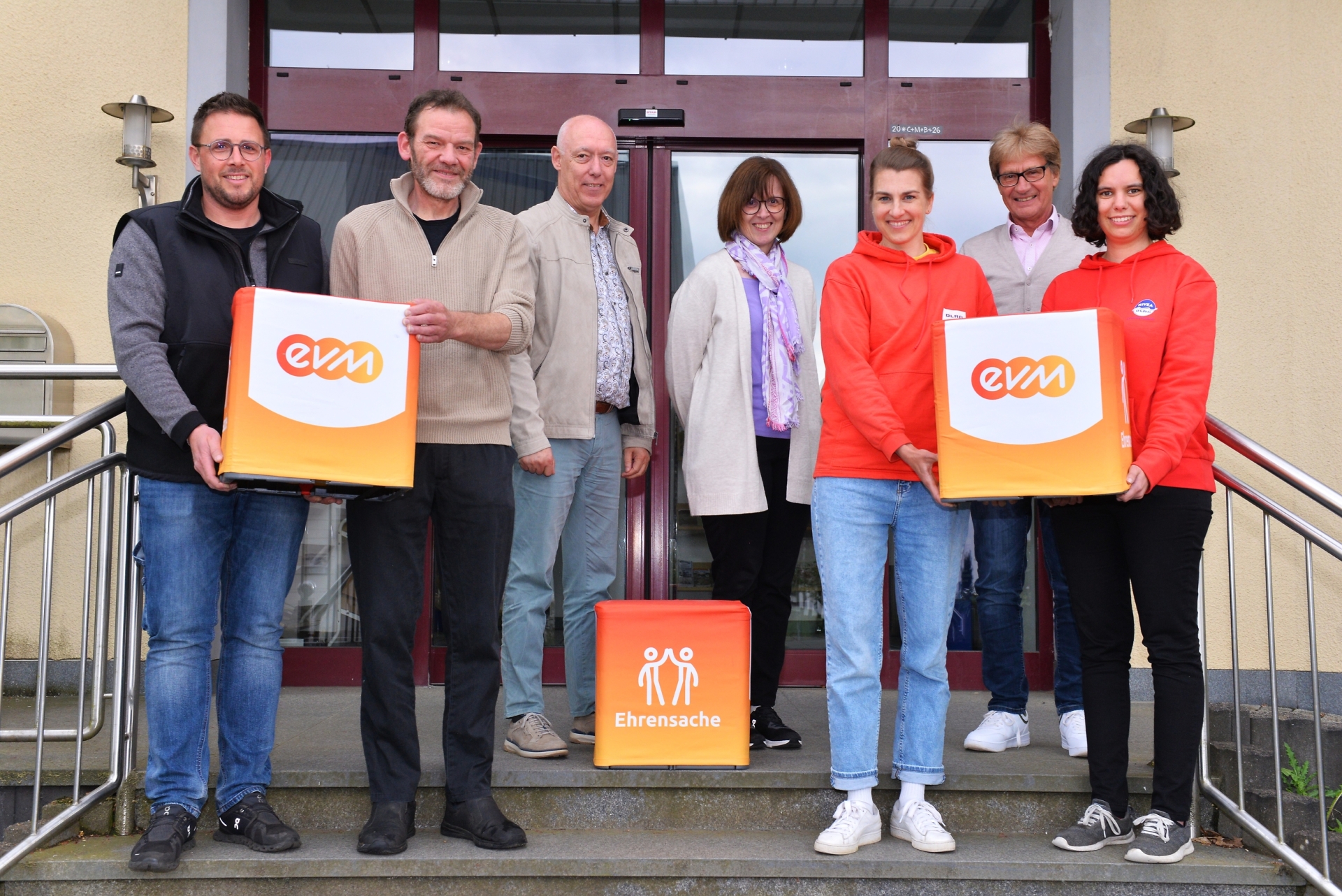 Berthold Nick von der evm und Bürgermeister Johannes Saxler stehen mit Vertreterinnen und Vertretern des Angelvereins Gunderath und der DLRG Ortsgruppe Kelberg vor einem Gebäude und halten „evm‑Ehrensache“-Spendenboxen bei der Übergabe.
