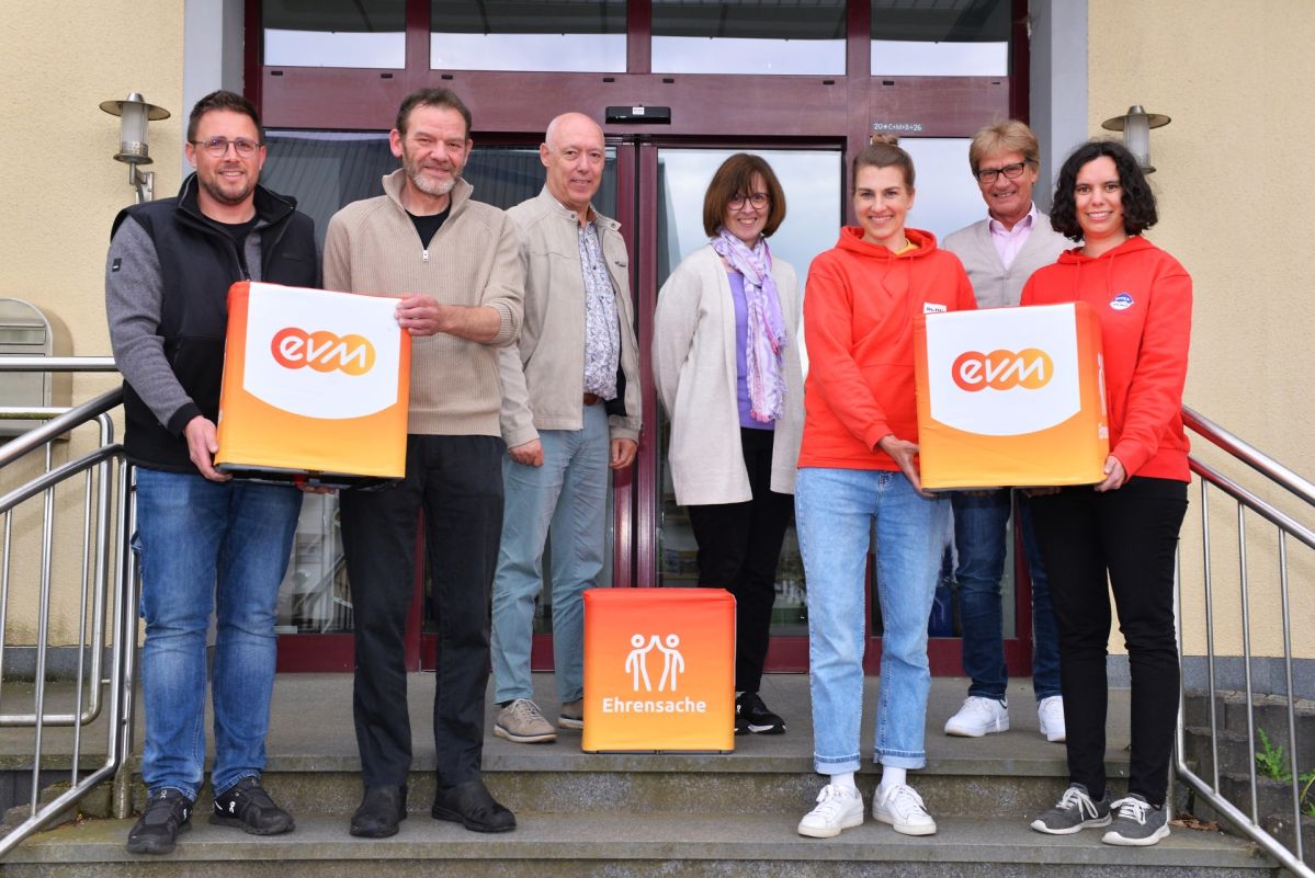 Berthold Nick von der evm und Bürgermeister Johannes Saxler stehen mit Vertreterinnen und Vertretern des Angelvereins Gunderath und der DLRG Ortsgruppe Kelberg vor einem Gebäude und halten „evm‑Ehrensache“-Spendenboxen bei der Übergabe.