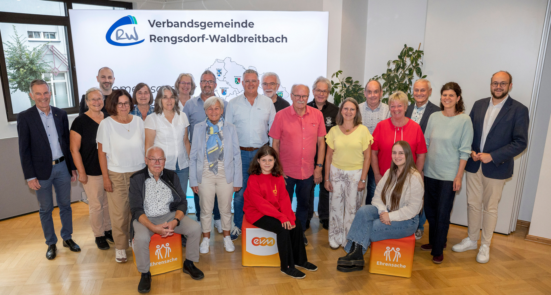 Vertreterinnen und Vertreter von elf Gesangsvereinen und Gesangsgruppen sowie zwei Kinderchören aus der Verbandsgemeinde Rengsdorf-Waldbreitbach stehen gemeinsam für ein Gruppenfoto vor einer Bannerwand der Verbandsgemeinde. Im Vordergrund sitzen drei Personen auf „evm‑Ehrensache“-Spendenboxen. Mit dabei sind Norbert Rausch von der evm und Bürgermeister Pierre Fischer, die die Spende aus dem Programm „evm‑Ehrensache“ an die begünstigten Vereine übergeben haben.