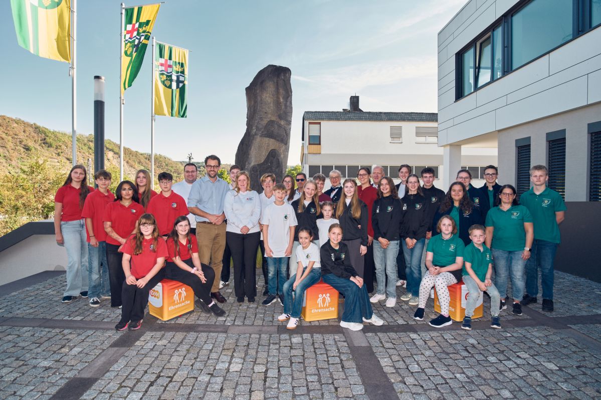 Vertreterinnen und Vertreter der DLRG Ortsgruppe Untermosel e.V. sowie der Jugendrotkreuz-Abteilungen des DRK Brodenbach/Oberfell e.V., DRK Waldesch e.V. und DRK Rhens e.V. stehen gemeinsam mit Hubertus Hacke-Dietze von der evm und Bürgermeisterin Kathrin Laymann vor dem Rathaus der Verbandsgemeinde Rhein-Mosel. Im Vordergrund sind „evm‑Ehrensache“-Spendenboxen zu sehen, die symbolisch für die übergebenen Fördergelder stehen.
