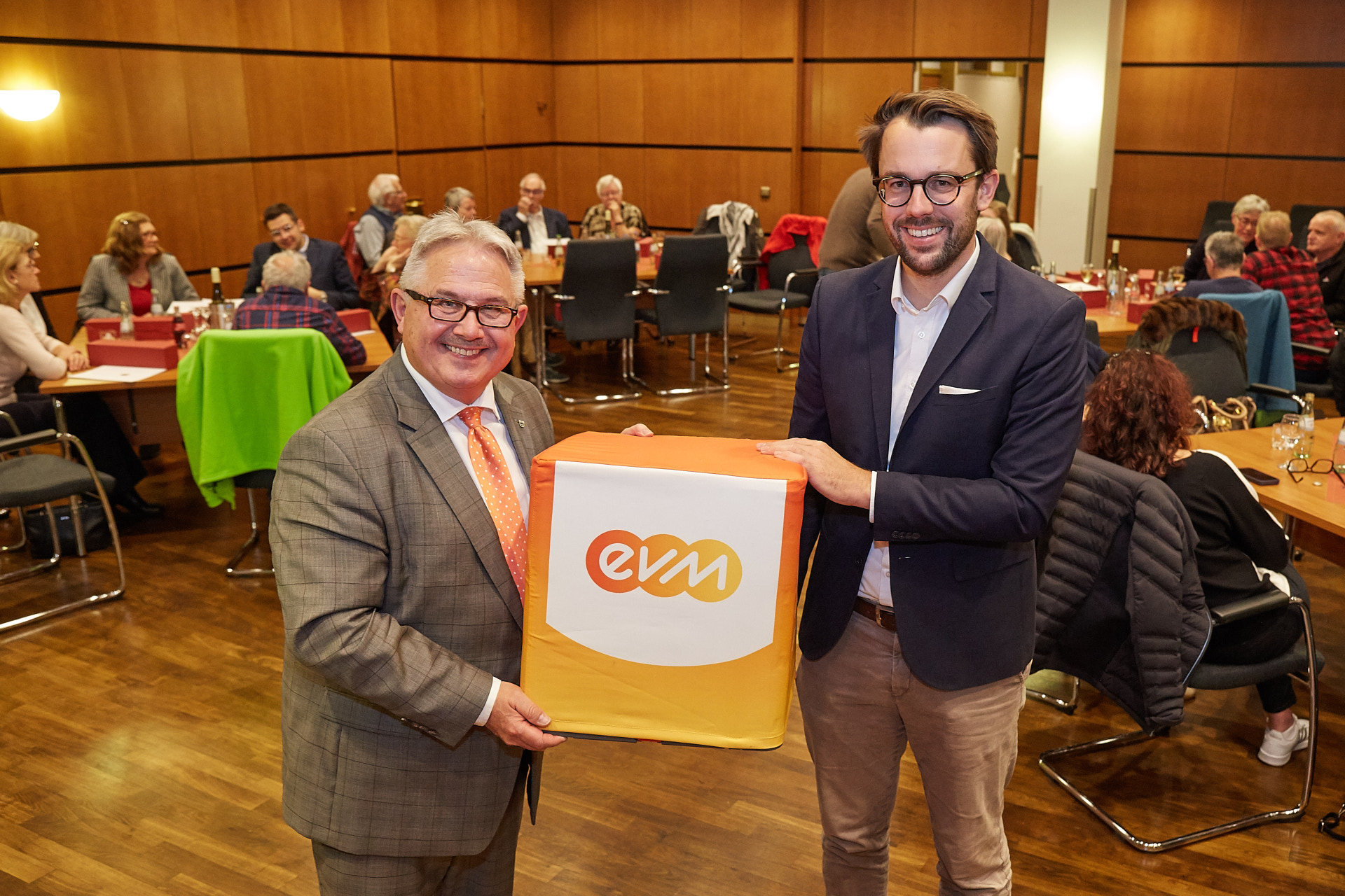 Zwei Männer halten einen großen, orangenen Würfel mit der Aufschrift "evm".