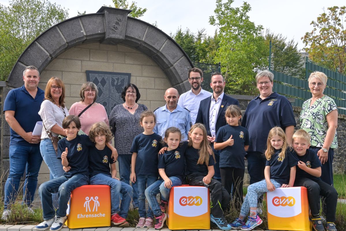 Ehrensache Vordereifel Mehrere Erwachsene und Kinder der Bambini-Feuerwehr Luxem sowie des Seniorenteams der Ortsgemeinde Reudelsterz stehen vor einem steinernen Ehrenmal. Im Vordergrund sitzen und stehen Kinder in einheitlichen T‑Shirts neben „evm‑Ehrensache“-Spendenboxen. Hubertus Hacke-Dietze von der evm und Bürgermeister Timo Kanzinger präsentieren gemeinsam mit den Vereinsvertretungen die Spende für die beiden Projekte.