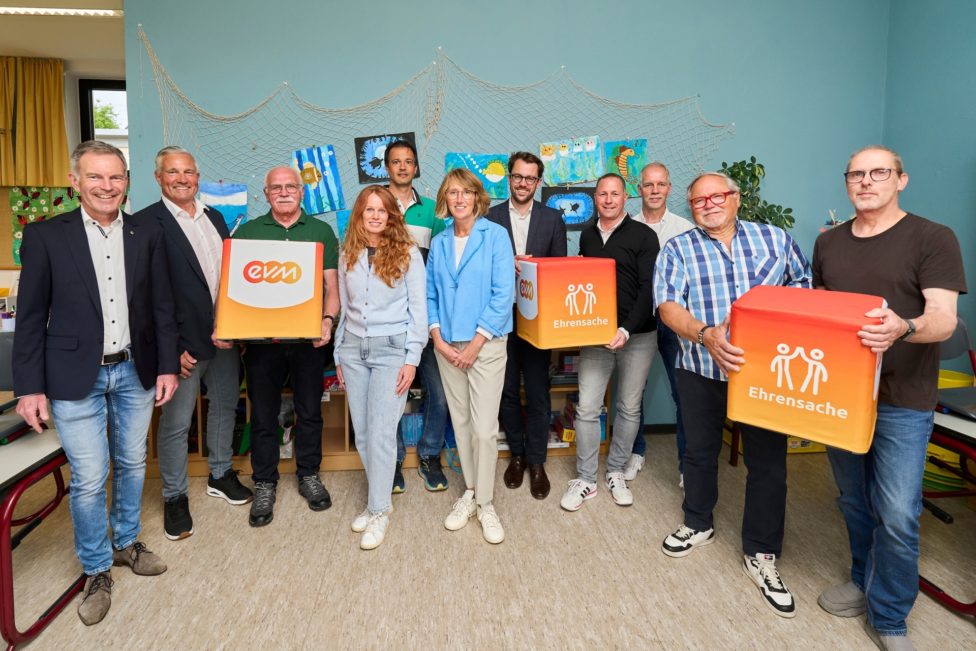 Norbert Rausch von der evm und Bürgermeister Klaus Lütkefedder stehen mit Vertreterinnen und Vertretern mehrerer Vereine in einem Raum mit Kinderbildern an der Wand und halten „evm‑Ehrensache“-Spendenboxen bei der Übergabe.