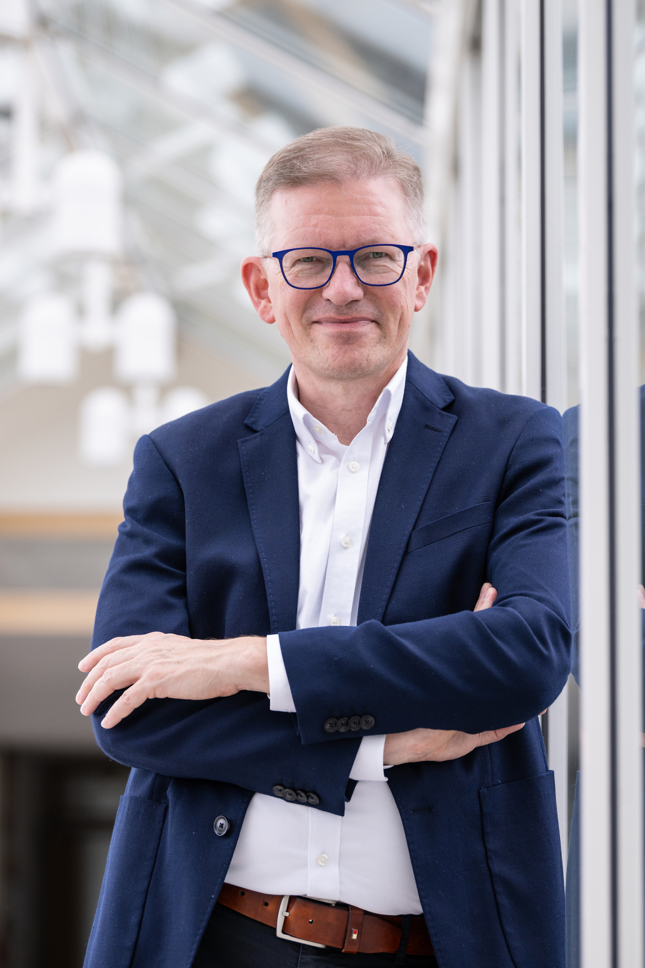 Marcel Peerenboom Marcelo Peerenboom steht mit verschränkten Armen in einem hellen, modernen Gebäude vor einer Glasfront. Der Unternehmenssprecher der evm ist Gastgeber des neuen Dialogformats „evm‑Energietreff“, bei dem Bürgerinnen und Bürger Fragen rund um Energie und die evm‑Gruppe stellen können.