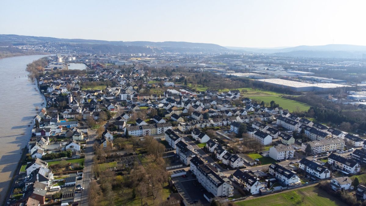 Luftaufnahme von Koblenz-Kesselheim am Rhein mit dicht bebauten Wohngebieten, Grünflächen und Gewerbearealen. Links verläuft der breite Fluss, während sich im Hintergrund Hügel und ein weitläufiges Stadtgebiet erstrecken.