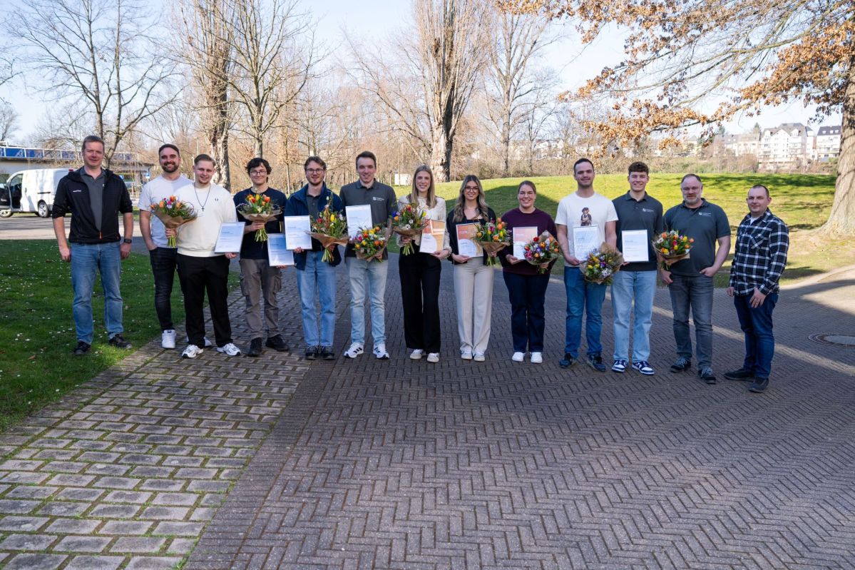 Ausbilder der evm‑Gruppe stehen im Freien mit elf frisch ausgebildeten Nachwuchskräften, die ihre Abschlusszeugnisse und Blumensträuße zum erfolgreichen Ausbildungsabschluss in den Händen halten.