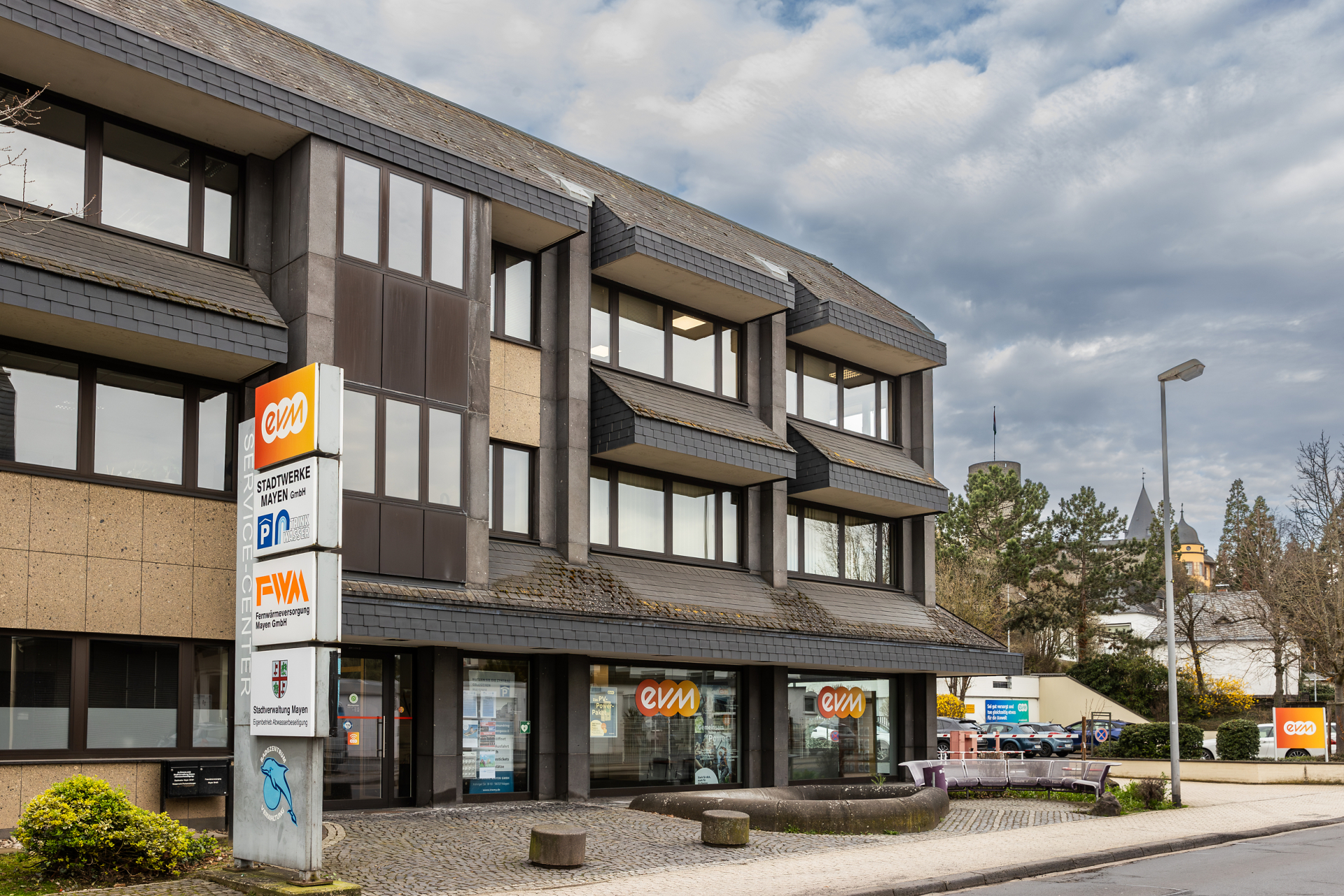 Das evm‑Kundenzentrum in Mayen befindet sich in einem mehrgeschossigen Bürogebäude an einer Straße. Vor dem Eingang stehen ein Hinweisschild mit den Logos von evm, den Stadtwerken Mayen und der Fernwärmeversorgung Mayen sowie Sitzgelegenheiten.
