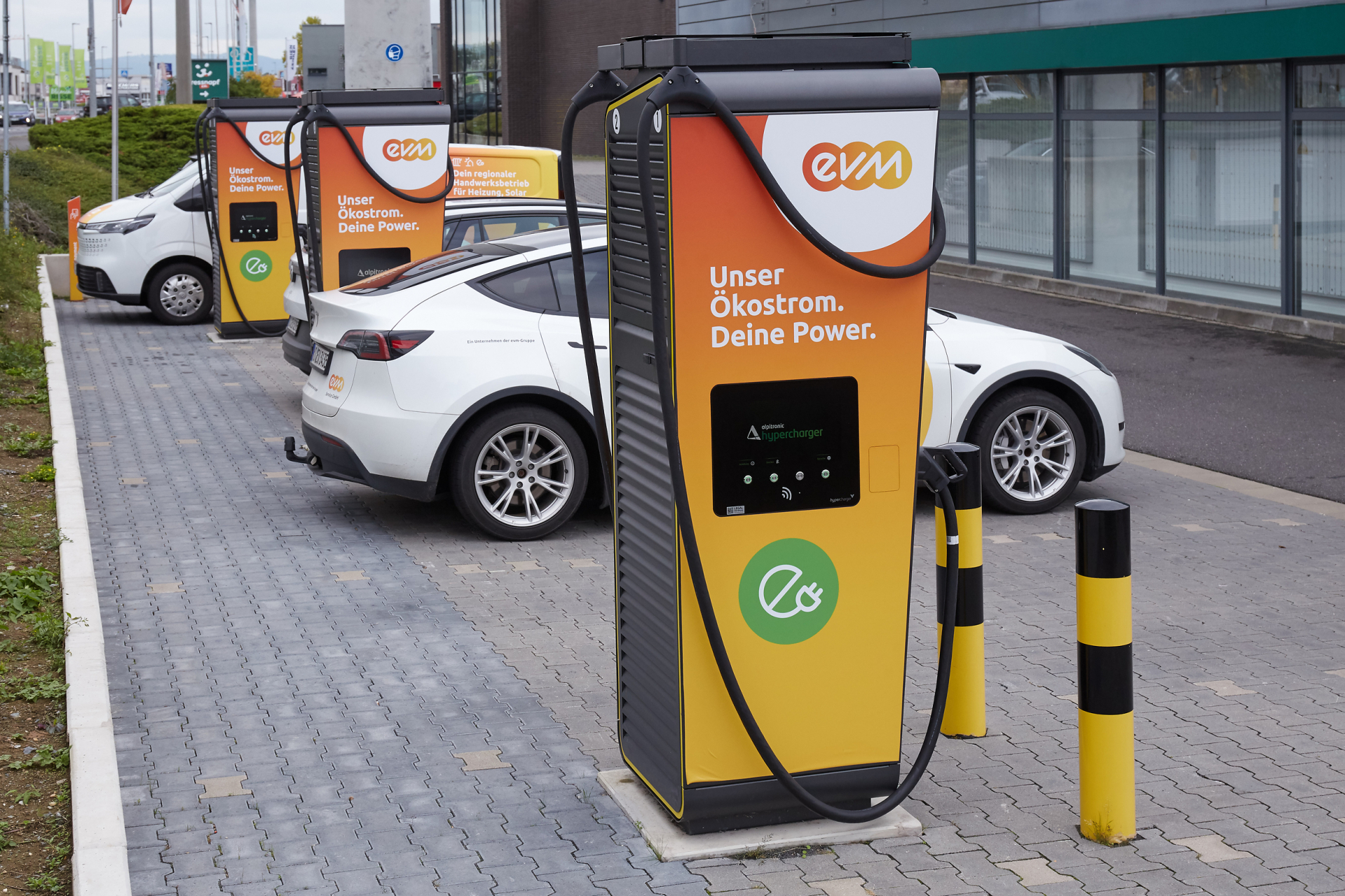 Mehrere evm‑Schnellladesäulen stehen auf einem Parkplatz vor einem Gewerbegebäude in Mülheim‑Kärlich. Neben den Ladesäulen sind Elektrofahrzeuge geparkt. An diesem Standort können E‑Autos an DC‑Ladepunkten mit bis zu 200 kW Leistung geladen werden.