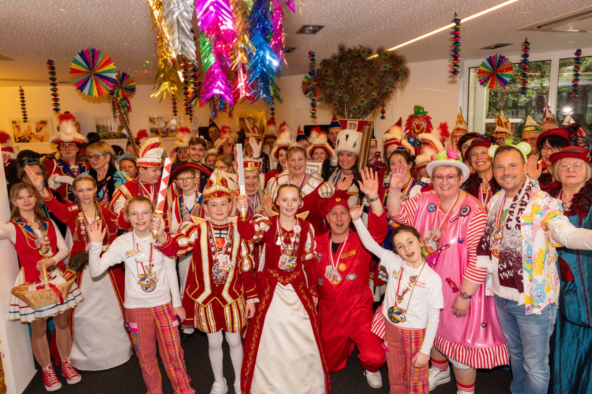 Alaaf am Linzer Netzstandort Große Gruppe von Karnevalistinnen und Karnevalisten in farbenfrohen Kostümen beim Prinzenempfang der evm‑Gruppe in Linz; darunter die Tollitätenpaare aus Linz und Kripp sowie das Kripper Dreigestirn, umgeben von bunt geschmücktem Raum mit karnevalistischen Dekorationen.
