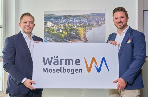 Norman Spaniol und Christopher Dold stehen in einem Büro und halten eine Tafel mit dem Logo der neuen Wärme Moselbogen GmbH. Die beiden Geschäftsführer präsentieren damit die gemeinsame Gesellschaft von evm und Iqony, die den Ausbau und die Weiterentwicklung der Wärmeversorgung im Koblenzer Rauental vorantreiben soll.