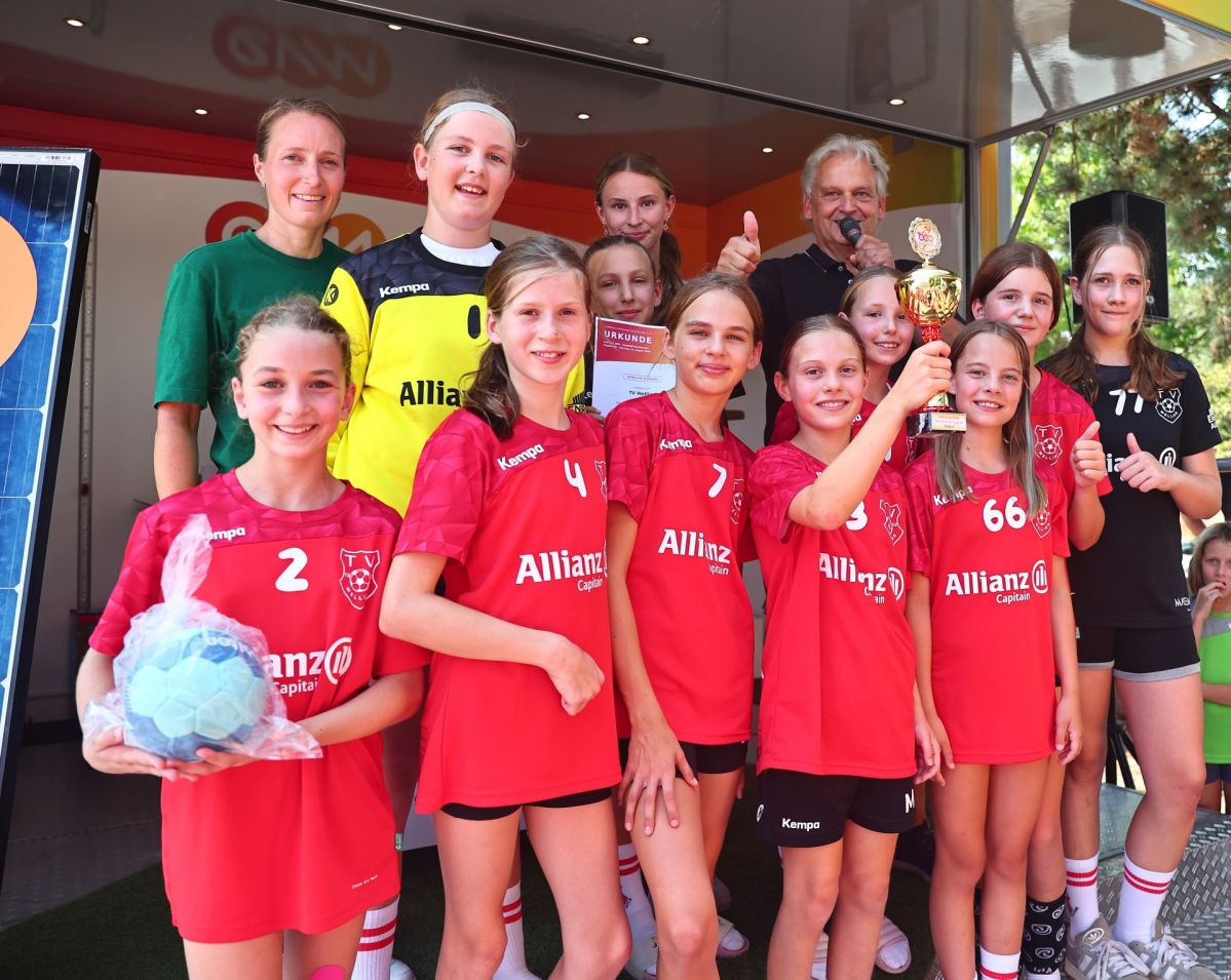 Die Spielerinnen des TV Welling II stehen gemeinsam mit ihren Trainerinnen und Trainern auf der Bühne des evm‑Infomobils. Eine Spielerin hält den Siegerpokal hoch, während eine weitere die Urkunde für den ersten Platz präsentiert.