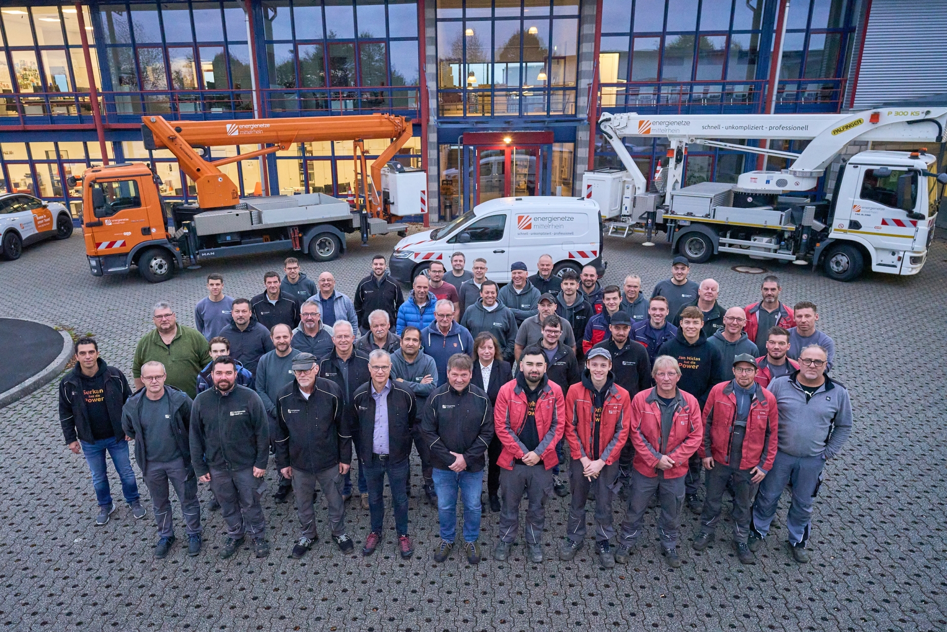 Mitarbeitende der Energienetze Mittelrhein stehen vor dem Betriebsgebäude des Standorts Hahn am See für ein Gruppenfoto. Im Hintergrund sind mehrere Servicefahrzeuge und Hubarbeitsbühnen des Netzbetreibers zu sehen. Das Team betreut von hier aus die Strom- und Gasversorgung in großen Teilen des Westerwalds und unterstützt die Umsetzung der Energiewende in der Region.
