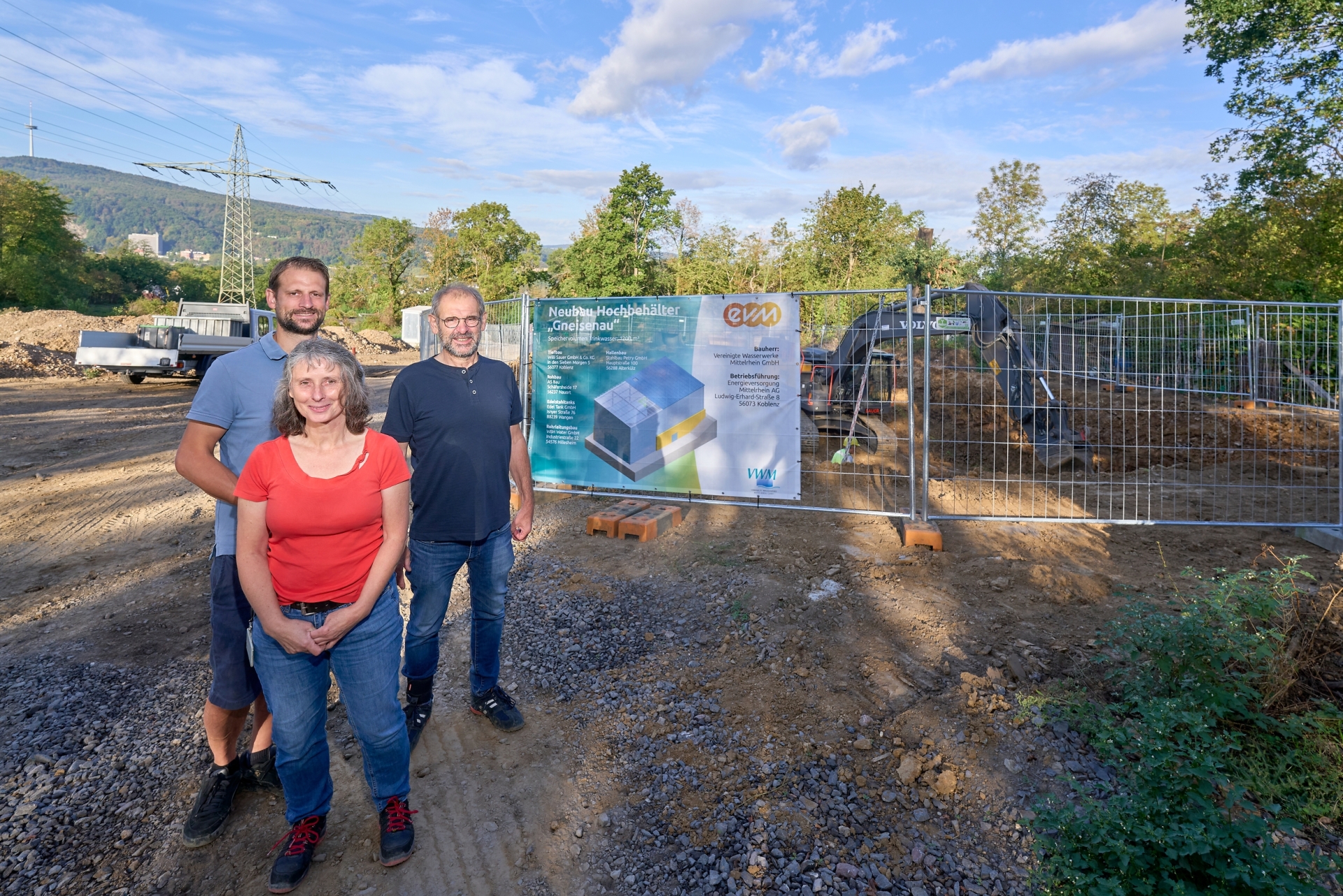 Viola Pöllmann-Kux von der Energieversorgung Mittelrhein (evm) sowie Maximilian Sauer und Paul Sauer von der Sauer Bauunternehmung stehen vor der Baustelle des neuen Hochbehälters Gneisenau. Im Hintergrund sind das Baufeld und ein Bagger bei den laufenden Tiefbauarbeiten zu sehen.