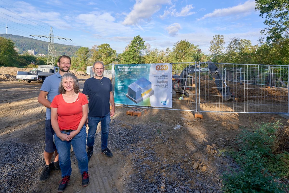 Neubau Hochbehälter „Gneisenau“ Koblenz-Horchheim Viola Pöllmann-Kux von der Energieversorgung Mittelrhein (evm) sowie Maximilian Sauer und Paul Sauer von der Sauer Bauunternehmung stehen vor der Baustelle des neuen Hochbehälters Gneisenau. Im Hintergrund sind das Baufeld und ein Bagger bei den laufenden Tiefbauarbeiten zu sehen.