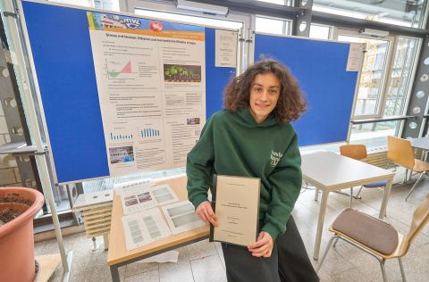 Jugend forscht 2026 - Regionalwettbewerb Koblenz Eine teilnehmende Person des Wettbewerbs „Jugend forscht“ steht vor einem Präsentationsstand und hält eine Projektmappe in den Händen. Hinter ihr befindet sich ein großes wissenschaftliches Poster zum Thema „Hormetische Effekte mit Hefe“, das Diagramme, Versuchsbeschreibungen und Forschungsergebnisse zeigt. Auf dem Tisch vor dem Poster liegen weitere Unterlagen und Ausdrucke, die zum Forschungsprojekt gehören.