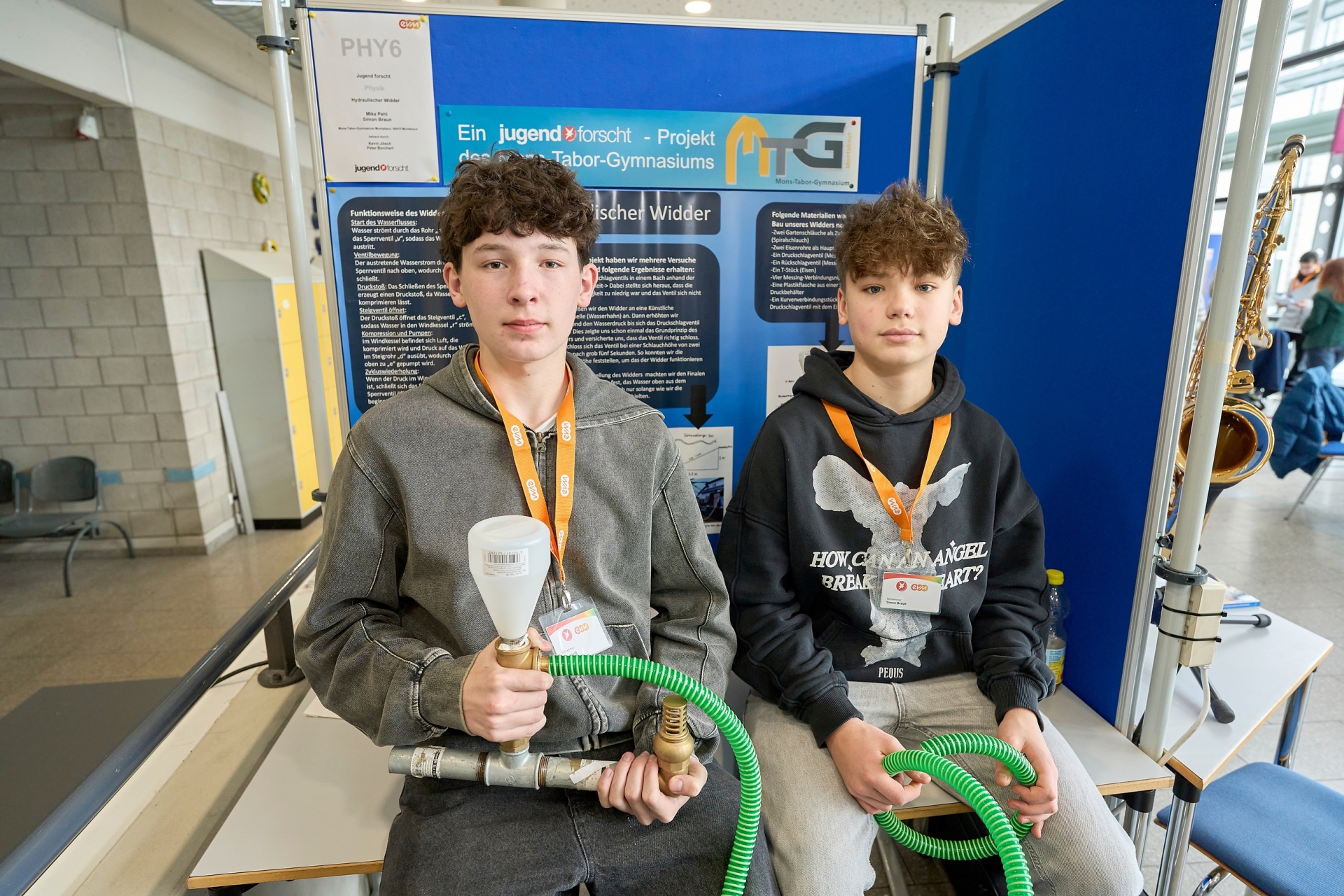 Zwei Teilnehmende präsentieren am „Jugend forscht“-Stand ihr Projekt zum hydraulischen Widder und halten dafür einen aufgebauten Versuchsaufbau mit Rohren und Ventilen in den Händen.