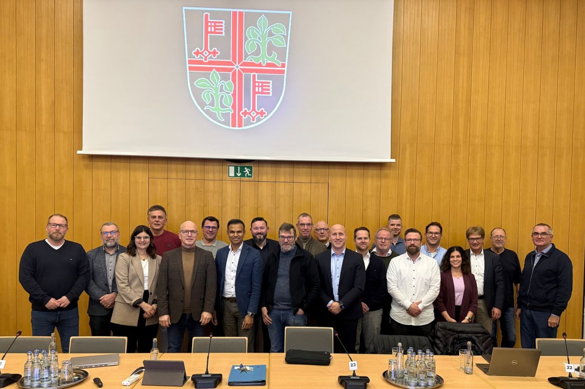 Gruppenfoto von Teilnehmenden in einem Sitzungssaal des Mayener Rathauses. Die Personen stehen vor einer Holzvertäfelung und unter einer Projektionsfläche, auf der ein farbiges Wappen zu sehen ist. Im Vordergrund sind Konferenztische mit Laptops, Mikrofonen und Getränken zu erkennen.