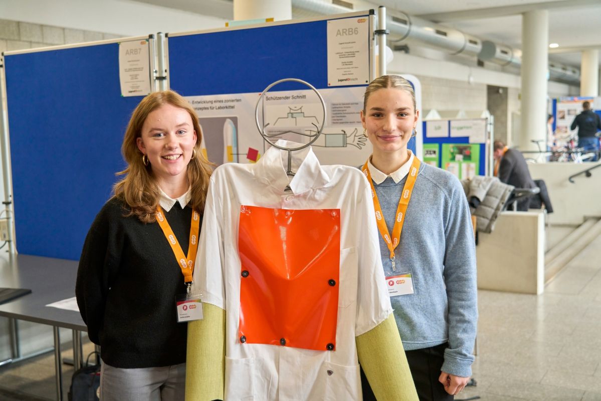 An einem „Jugend forscht“-Stand wird ein modifizierter Laborkittel mit großem orangefarbenem Schutzpanel präsentiert, begleitet von einer Stellwand mit Projektinformationen zu Sicherheitskleidung.