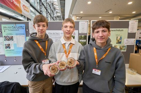 Drei Teilnehmende von „Jugend forscht“ zeigen am Präsentationsstand ihr Projekt, indem sie mehrere Petrischalen mit Pilzkulturen in den Händen halten. Hinter ihnen befinden sich Poster mit Informationen zu ihrem Forschungsansatz.