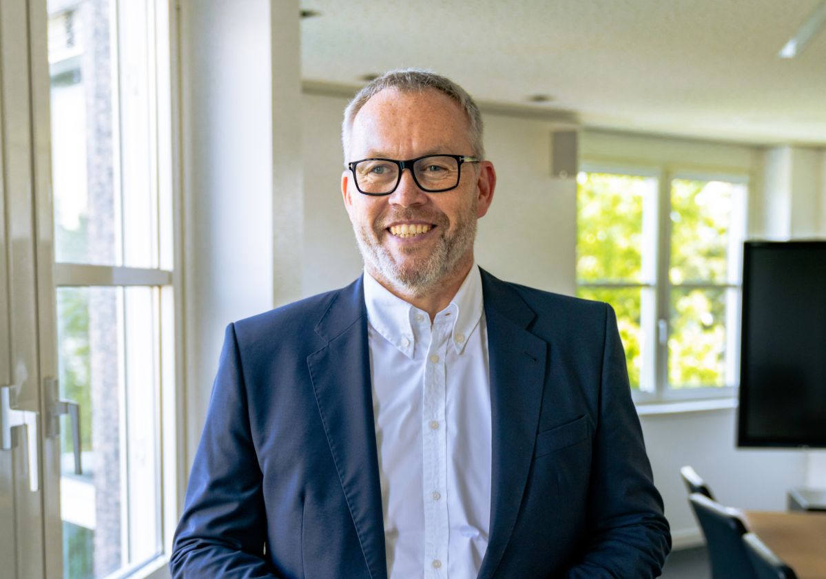 Der neue Vertriebschef der evm, Oliver Felthaus,  steht in einem dunkelblauem Anzug und weißem Hemd in einem hellen Büro und hält die Hände locker vor dem Oberkörper.