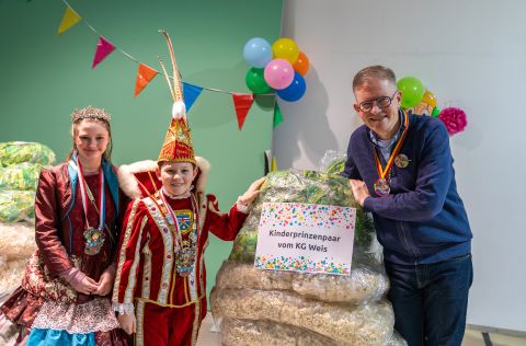 Palettenparty Kinderprinzenpaar der KG Weis steht mit einem Vertreter der evm neben einer Palette voller Wurfmaterial, dekoriert mit bunten Girlanden.