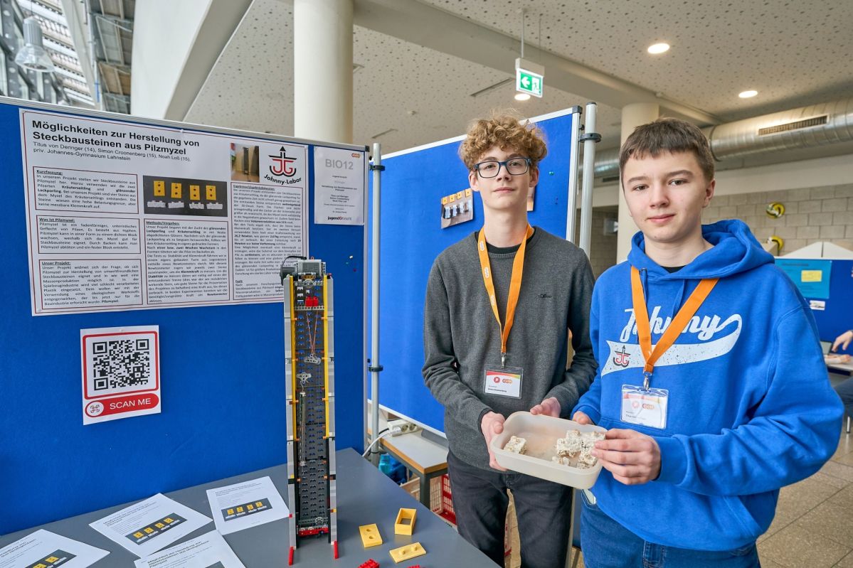 Zwei Teilnehmende präsentieren ihr „Jugend forscht“-Projekt zur Herstellung von Steckbausteinen aus Pilzmyzel an einem Ausstellungsstand mit Poster, Modellaufbau und Beispielbausteinen.