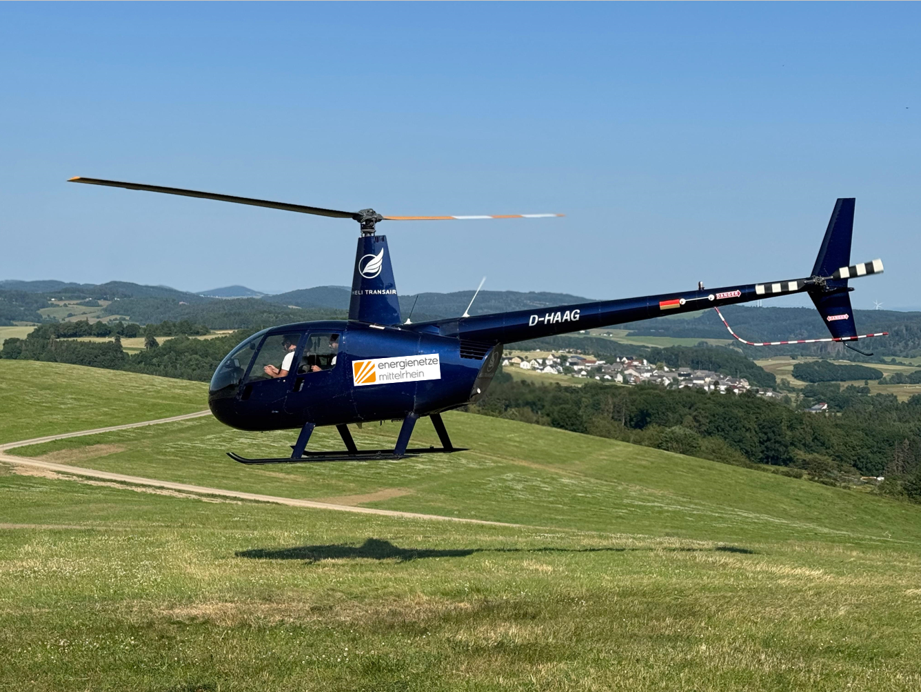 Ein Helikopter der Energienetze Mittelrhein fliegt in geringer Höhe über eine Wiese und inspeziert Stromleitungen im ländlichen Gebiet zwischen Eifel und Westerwald.