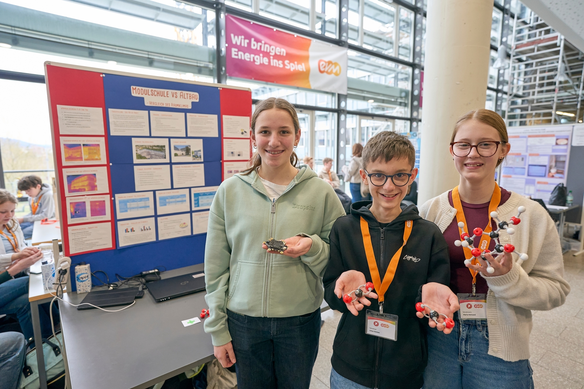 Drei Teilnehmende von „Jugend forscht“ präsentieren an ihrem Stand ein Projekt zum Vergleich von Holz- und Filterbaustoffen und halten Modellmoleküle sowie Versuchsmaterialien in den Händen; hinter ihnen steht eine Stellwand mit Informationspostern.