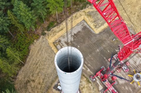 Turmbau Windpark Schneifelhöhe Die Luftaufnahme zeigt die Baustelle einer Windenergieanlage im Windpark Schneifelhöhe. Ein großer roter Raupenkran montiert weitere Turmsegmente auf einem bereits weit fortgeschrittenen Betonturm. Rund um den Standort sind Baustellenflächen, Maschinen und Waldflächen zu erkennen. Die Aufnahme dokumentiert den Baufortschritt der elf Windenergieanlagen auf der Schneifelhöhe.
