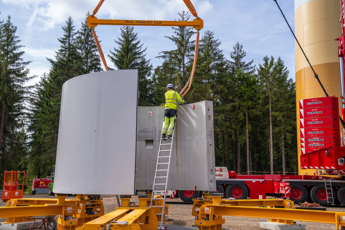 Turmbau Windpark Schneifelhöhe Ein Mitarbeitender begleitet die Montage eines Betonturmsegments für eine Windenergieanlage im Windpark Schneifelhöhe. Das Bauteil wird von einem Kran angehoben und über dem Turm positioniert. Im Hintergrund sind Kräne und Waldflächen der Baustelle zu sehen.