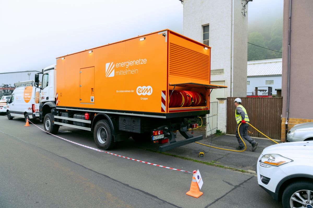 Selbstfahrer Das mobile Stromaggregat der Energienetze Mittelrhein steht am Straßenrand und versorgt über angeschlossene Kabel einen Netzabschnitt während Wartungsarbeiten. Das orangefarbene Fahrzeug mit integrierter Notstromtechnik ermöglicht eine unterbrechungsfreie Stromversorgung bei Arbeiten am Stromnetz.