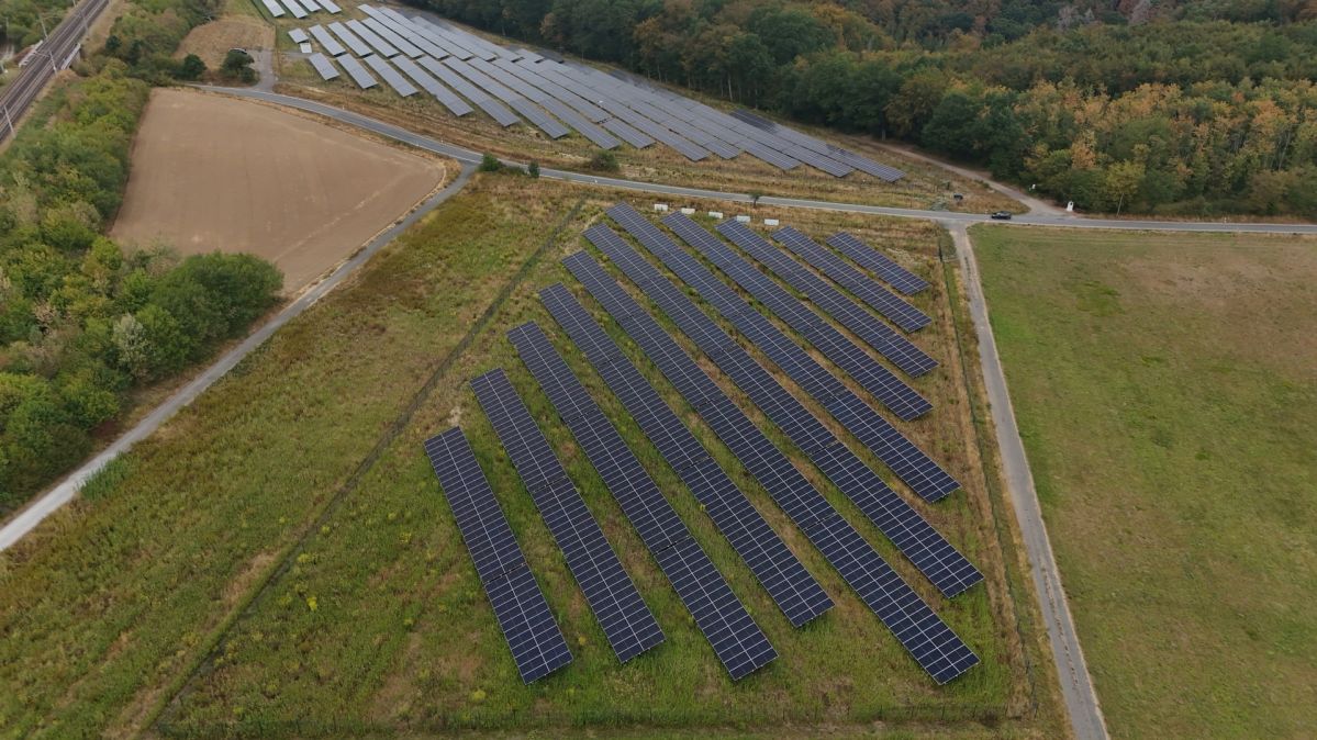 Solarpark Görgeshausen Das Luftbild zeigt den Solarpark Görgeshausen mit mehreren Reihen von Solarmodulen auf zwei eingezäunten Flächen zwischen Feldern, Wald und einer Bahnstrecke. Die Anlage wurde zuletzt um einen dritten Bauabschnitt erweitert und produziert heute so viel Strom aus erneuerbaren Energien, wie rund 3.900 Menschen im Jahr verbrauchen.