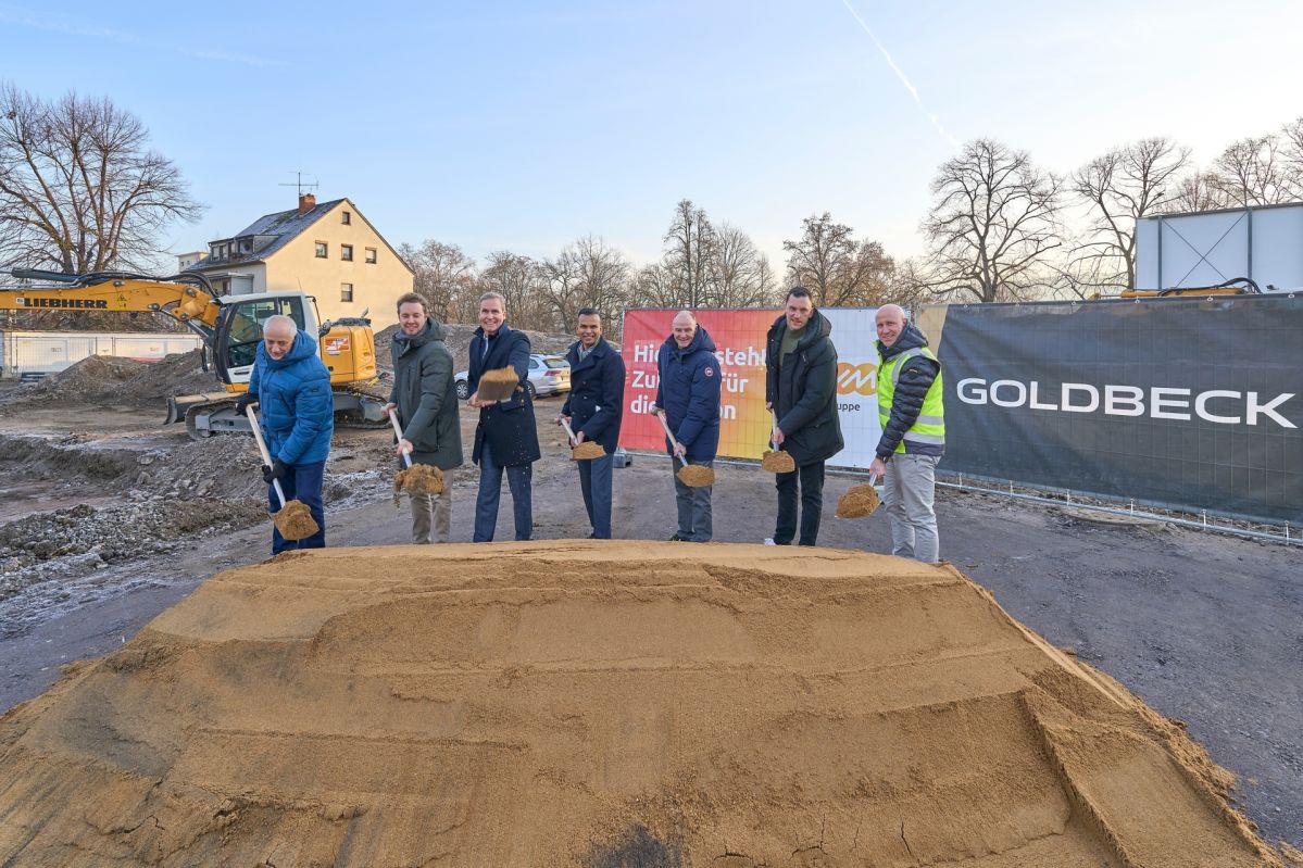 Personengruppe beim Spatenstich für den Neubau des evm‑Campus an der Schützenstraße in Koblenz, auf einer Baustelle mit Bagger, Bauzaun und Unternehmensbanner im Hintergrund.